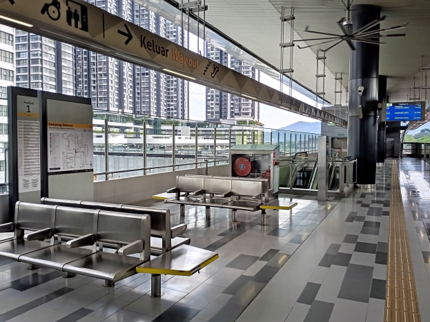 Leerer Flughafen Wartebereich mit mehreren Bänken, Informationsschildern, Säulen, Deckenventilator und Deckenbeleuchtung; Hintergrund zeigt Gebäude, Bäume, Wasser und einen klaren blauen Himmel.
