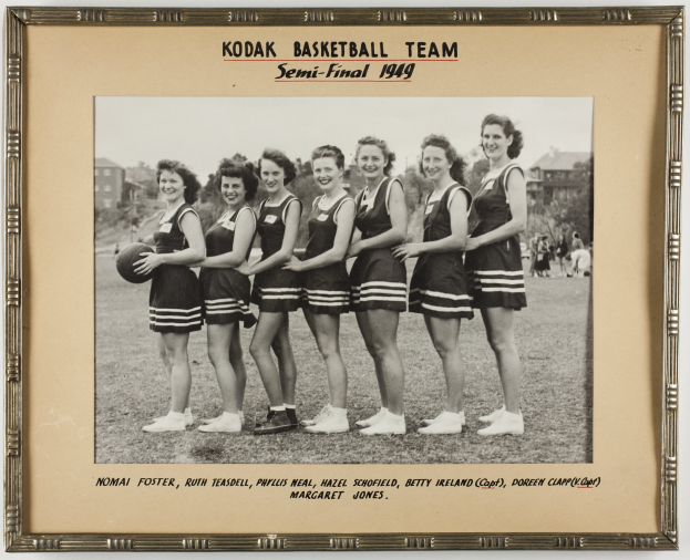 Schwarzes und weißes Foto einer Gruppe von Frauen der Kodak Basketball-Mannschaft aus dem Jahr 1949, die lächeln und einen Basketball halten, mit Menschen, Bäumen, Gebäuden und dem Himmel im Hintergrund.
