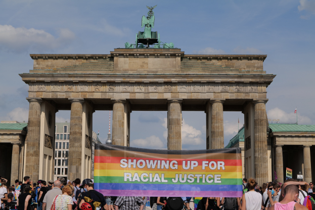 Eine Gruppe von Menschen hält eine "Racial Justice"-Schleife vor dem Brandenburger Tor in Berlin, mit den Säulen und der Statue des Tors im Hintergrund und Gebäuden und bewölktem Himmel.