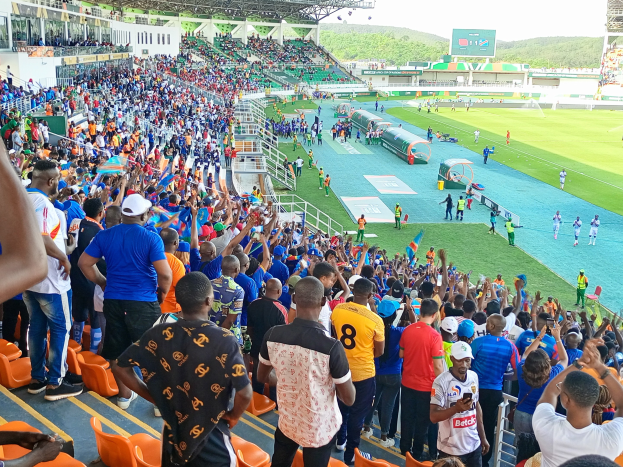 Eine große Menschenmenge versammelt sich vor einem eingezäunten Fußballfeld, einige sitzen auf Stühlen, andere stehen, unter einem klaren blauen Himmel mit Hügeln im Hintergrund und warten auf das Spiel der afrikanischen Nationenmeisterschaft 2019.