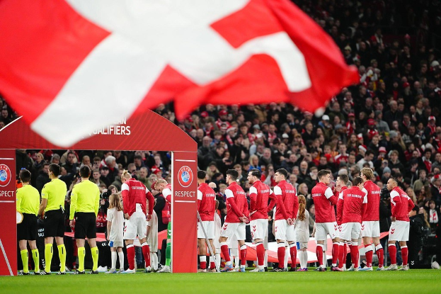 Eine Gruppe von Menschen auf einem Fußballfeld mit einer roten und weißen Fahne im Vordergrund, einem Bogen mit der Aufschrift "Bayern München vs. Bayern München Wetten & Vorschau" im Hintergrund und einer großen Menge im Stadion.