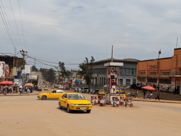 Ein gelbes Taxi fährt auf einer belebten Stadtstraße mit Gebäuden an den Seiten, während Fußgänger mit Regenschirmen auf dem Gehweg gehen, unter einem bewölkten Himmel, mit Strommasten und Kabeln im Hintergrund.
