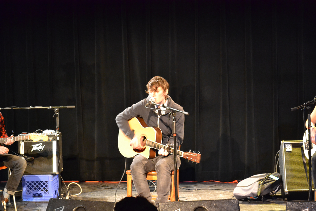 Eine Gruppe von Menschen, die vor einem Mikrofon Gitarre spielen, mit Lautsprechern, Gepäck und Vorhängen im Hintergrund.