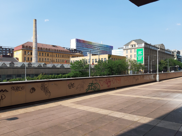 Stadtstraßenansicht mit Graffiti-beschmierten Wand, Bäumen, Straßenlaternen, einem Schild, Fahrzeugen, Gebäuden mit Fenstern, einem Schornstein und einem bewölkten Himmel.