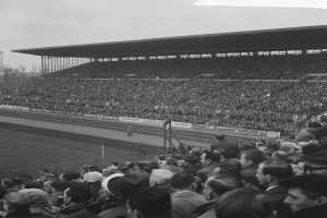 Schwarzes und weißes Foto eines vollen Fußballstadions mit Zuschauern, die ein Spiel verfolgen, mit Bannern, Pfählen, einer Hütte, Bäumen, einem Turm und einem bewölkten Himmel.
