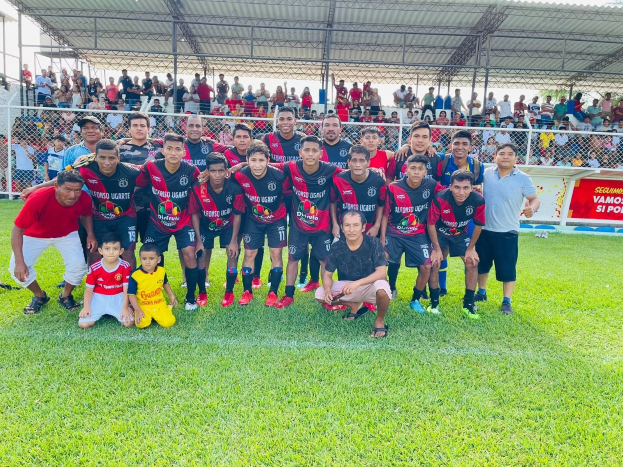 Mannergruppe feiert auf einem Fußballfeld mit einem Siegesschild, das "Fotos Ugarte U-17 vs U-18" lautet, unter einem bewölkten Himmel.