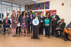 Eine Gruppe von Menschen steht und sitzt in einem Raum mit einem Podium, einem Mikrofon und einer Tafel mit der Aufschrift "Sancho City Councilman Joins City Council", mit Flaggen, einem Feuerlöscher und Deckenleuchten an der Rückwand.