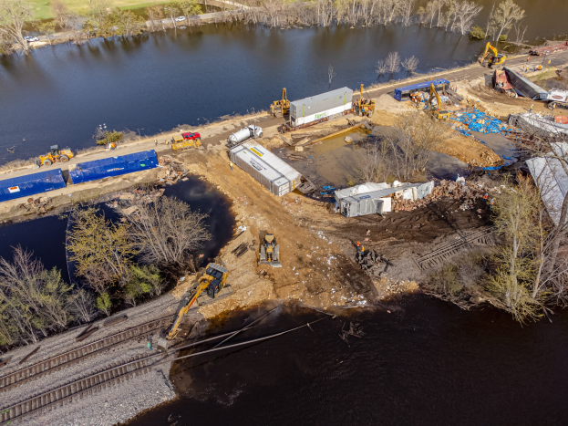Luftaufnahme einer Baustelle am Wasser mit Fahrzeugen, Bäumen, Gras und einer Brücke im Hintergrund, mit Geräten und Materialien, die auf ein Wasseraufbereitungsprojekt hinweisen.
