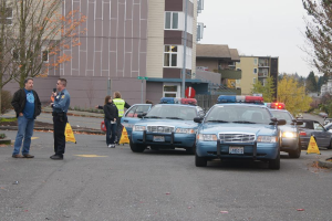 Autos auf einer Straße mit vier Menschen in der Nähe, Gebäude mit Fenstern im Hintergrund, Bäume und Warnbaken.