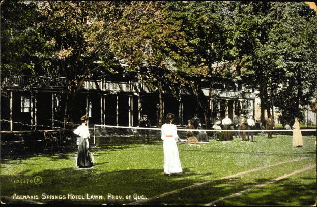 Altes Schwarz-Weiß-Foto einer Gruppe, die auf dem Rasen vor dem Abenakis Springs Hotel in Provo, Quebec, Tennis spielt, mit einem Netz, sitzenden Zuschauern, Bäumen und einem Gebäude im Hintergrund.