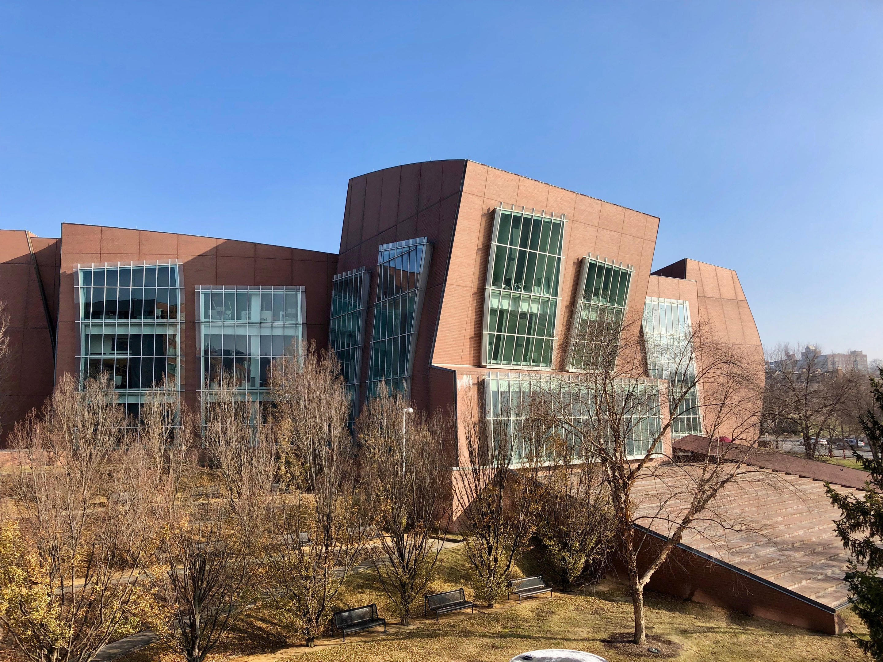 Großes Gebäude mit vielen Fenstern, umgeben von Bäumen, Bänken, Gras und einem Laternenmast, mit anderen Gebäuden und einem klaren blauen Himmel im Hintergrund, als der neue Campus der University of Illinois at Chicago identifiziert.