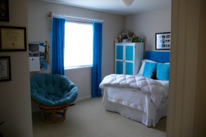 Ein Raum mit einem Bett, Stuhl, Fenster mit Vorhängen, Schrank, Foto Rahmen und einem gefliesten Boden.